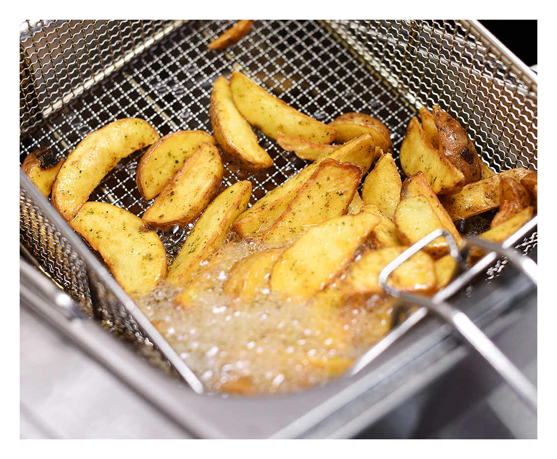 A deep fryer basket filled with golden-brown potato wedges sizzling in hot oil, with bubbles forming around them as they cook